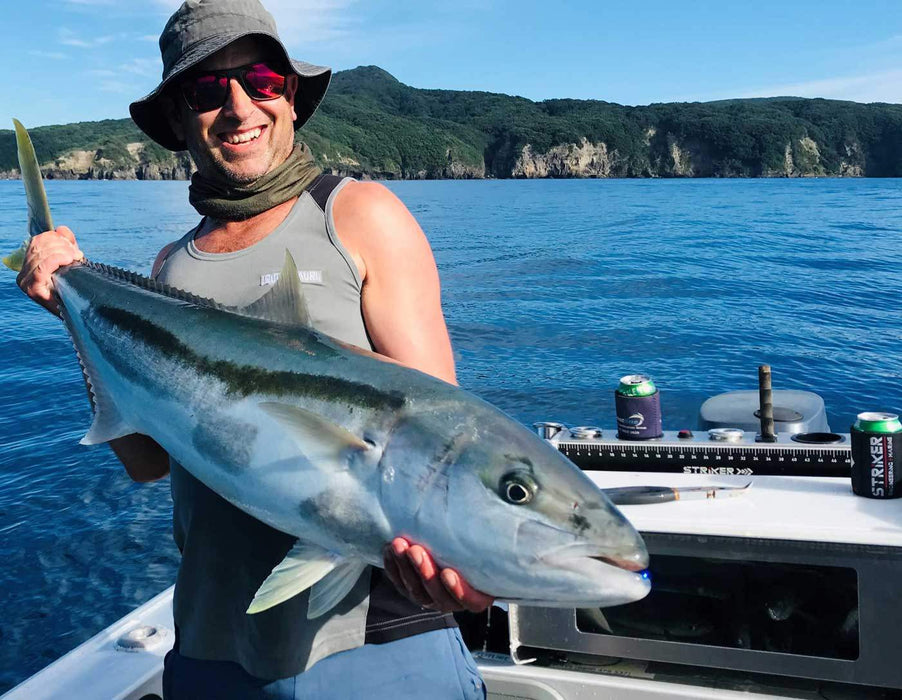 A NZ fisherman is holding a large fish while standing infront a Striker Marine bait board. The board has a cutting surface, a measuring ruler, and multiple rod holders. The fisherman is smiling and holding the fish up for the camera.