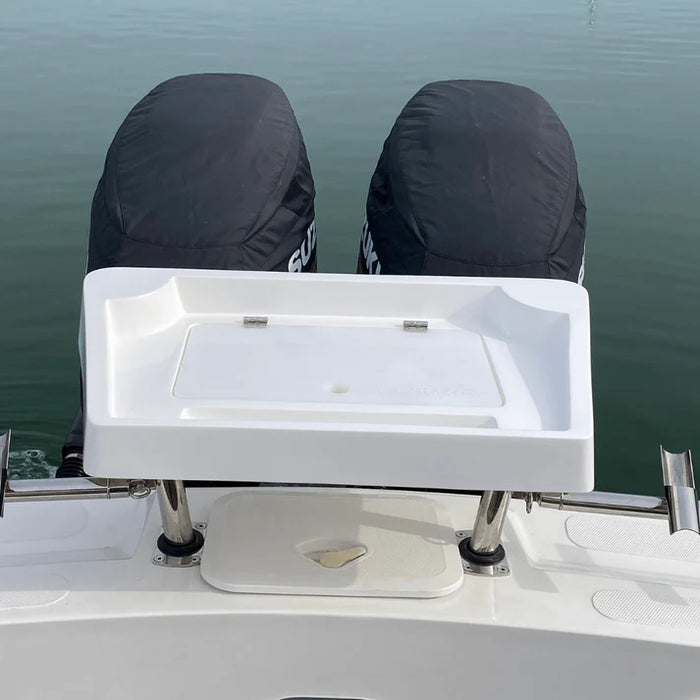 A white, rectangular fishing bait station mounted on the back of a boat with two outboard motors covered in black engine covers. The station has a hinged lid that is closed. There are two rod holders built into the back of the station, and a small cutting board is included.