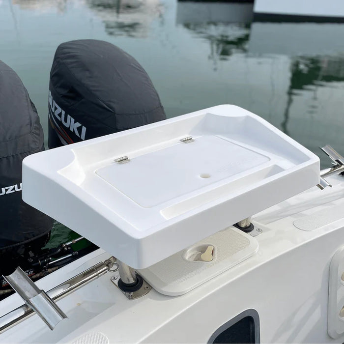 A white, rectangular fishing bait station mounted on the back of a boat with two outboard motors covered in black engine covers. The station has a hinged lid that is closed. There are two rod holders built into the back of the station, and a small cutting board is included.