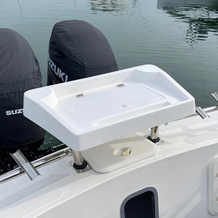 A white, rectangular fishing bait station mounted on the back of a boat with two outboard motors covered in black engine covers. The station has a hinged lid that is partially open, revealing a large, rectangular compartment with several smaller compartments around it. The "SUZUKI" logo is visible on one of the engine covers. There are two rod holders built into the back of the station, and a small cutting board is included.