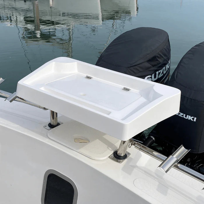 A white, rectangular fishing bait station mounted on the back of a boat with two outboard motors covered in black engine covers. The station has a hinged lid that is partially open, revealing a large, rectangular compartment with several smaller compartments around it. The "SUZUKI" logo is visible on one of the engine covers. There are two rod holders built into the back of the station, and a small cutting board is included.